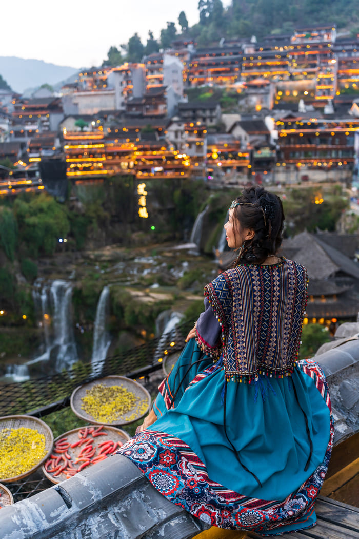 Persona con traje azul sentada de espaldas, mirando un pueblo de China iluminado por las cascadas de la montaña
